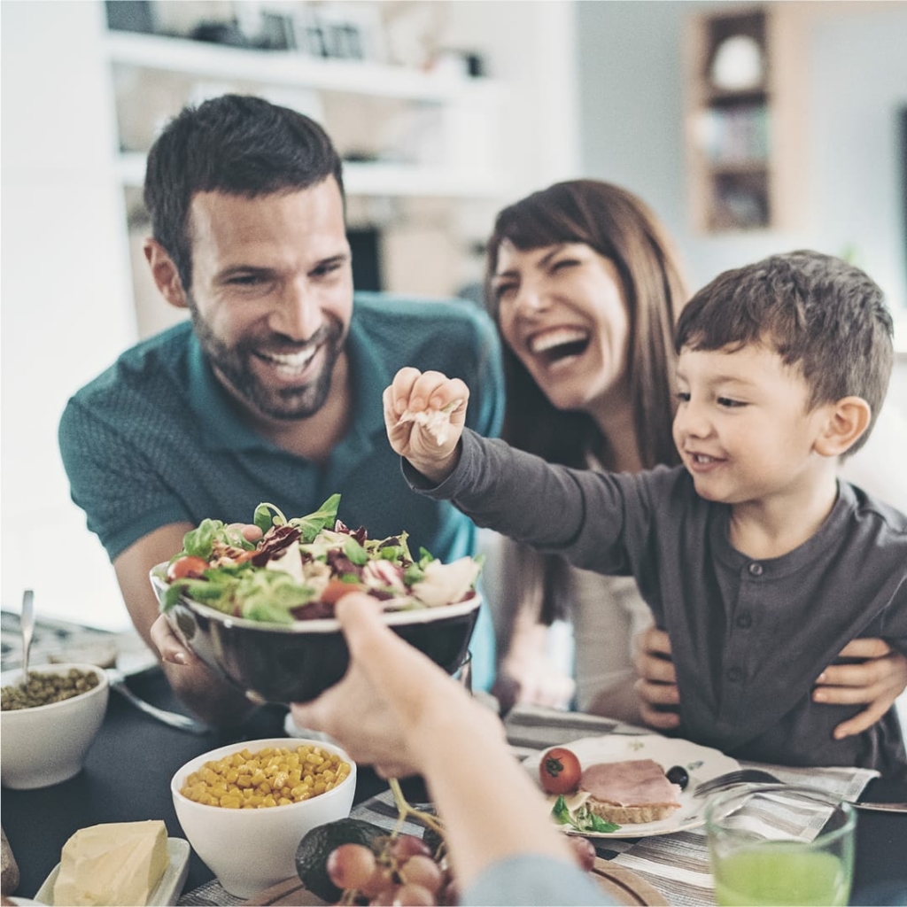 Lachende Familie sitzt gemeinsam am Esstisch; ein Kind greift nach einem Salat, der von einem Erwachsenen gehalten wird.