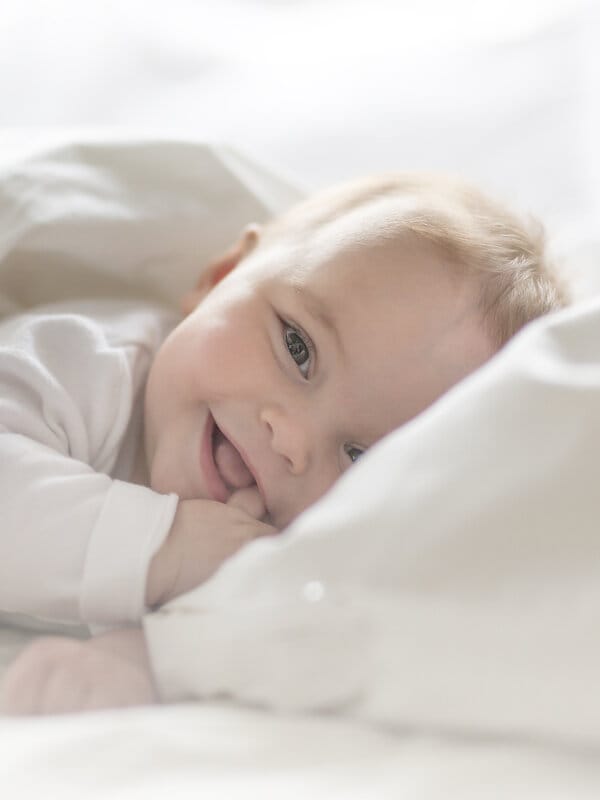 Lächelndes Baby unter weißer allergikerfreundlicher Bettwäsche.